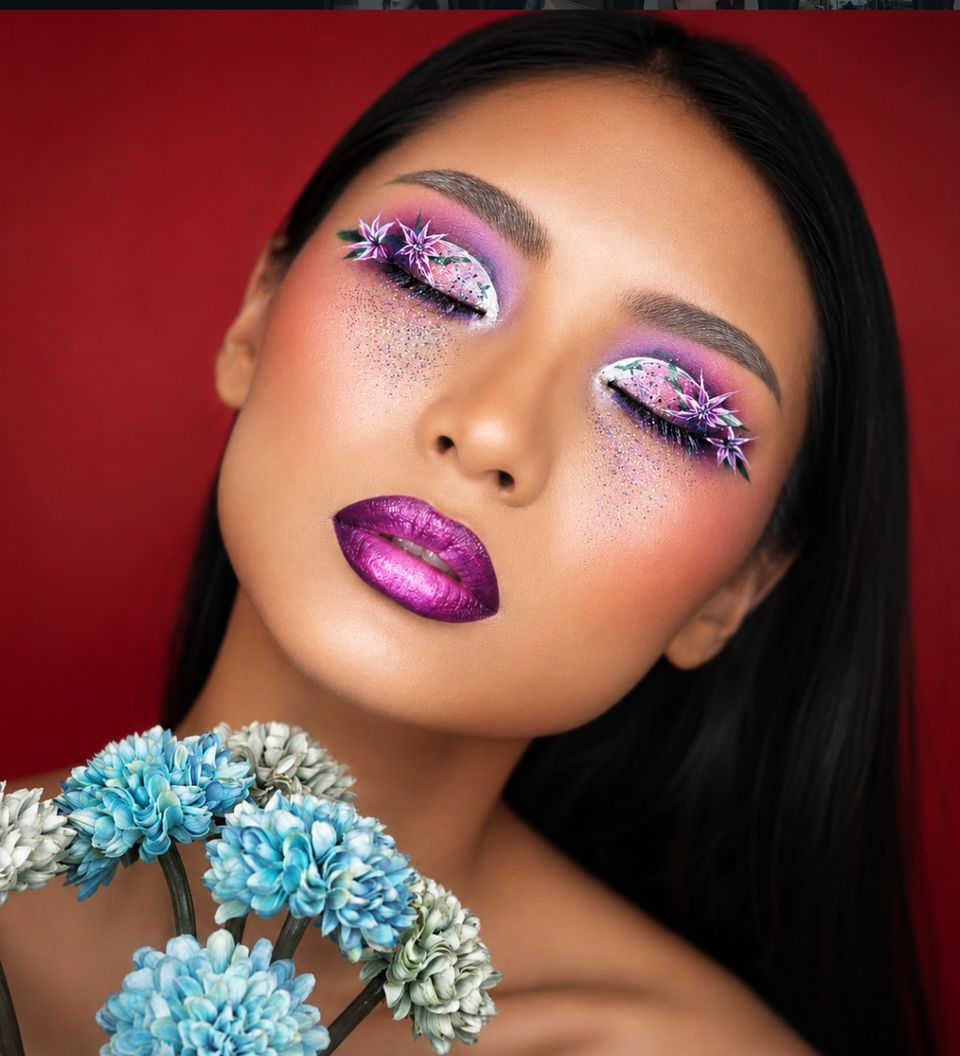 Model with iridescent magenta and lilac glitter eye makeup, rhinestone accents, holding pale blue chrysanthemums against a deep maroon backdrop