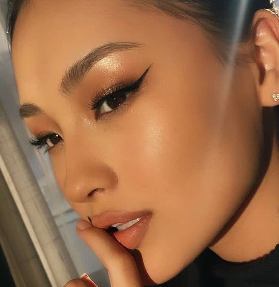 Close-up of glowing bronzed skin with sharp winged eyeliner, soft gold glitter eyelid, and glossy peach lip
