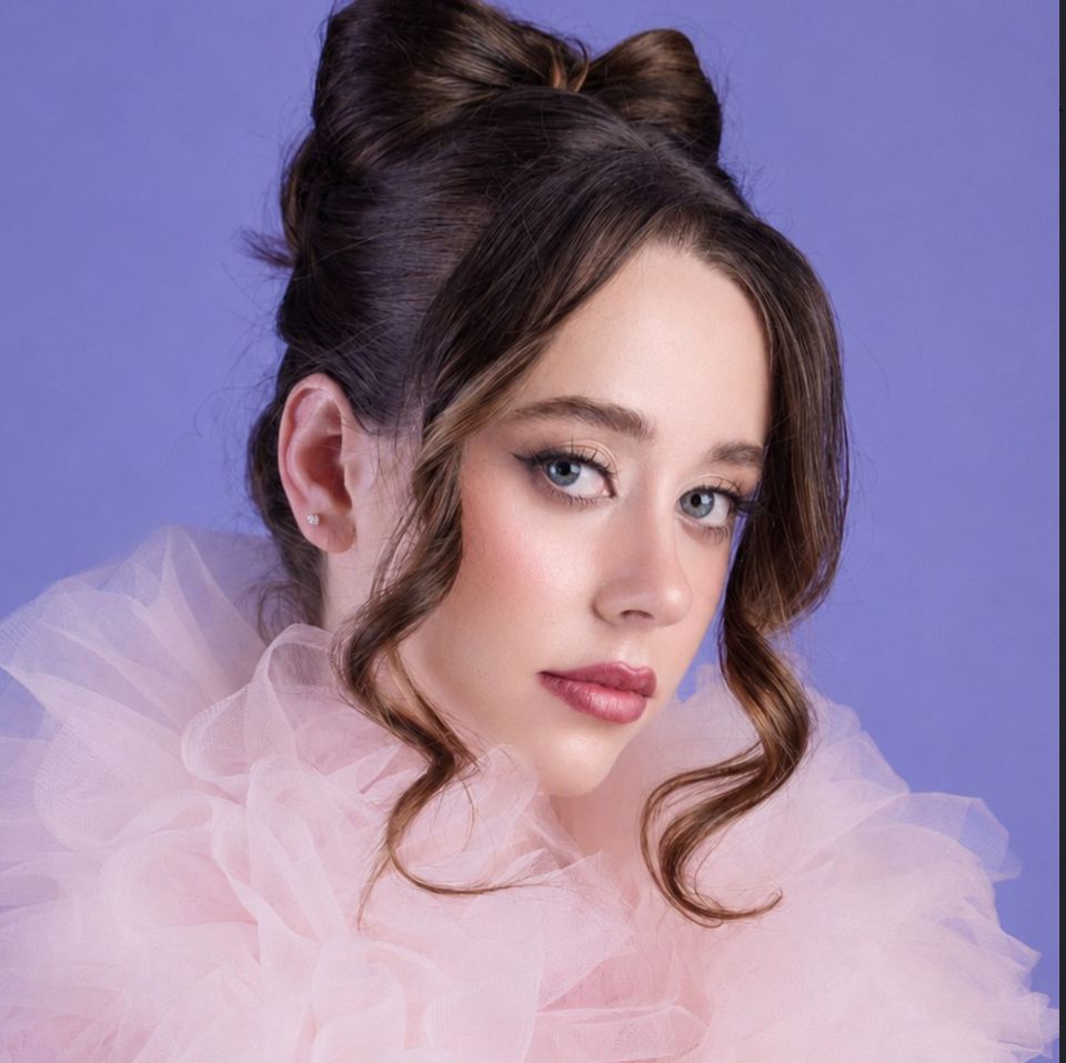 Model with a bow-shaped topknot hairstyle, blush-pink tulle collar, lilac backdrop, and soft pastel pink-lilac eye makeup