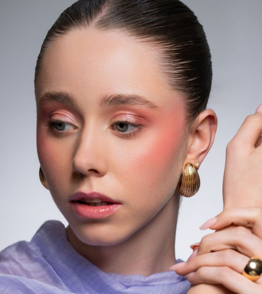 Soft minimal beauty look with coral-pink blush and eyeshadow, glossy nude-pink lip, and a gold shell earring
