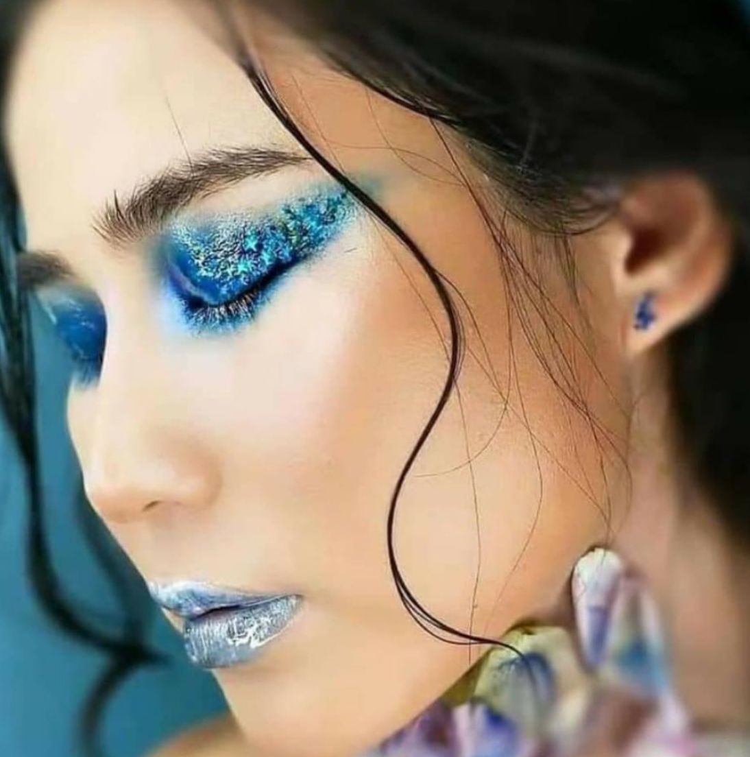Profile portrait with icy blue crystal and glitter eye makeup extending toward the temple, frosted blue lips, and a flower at the cheek