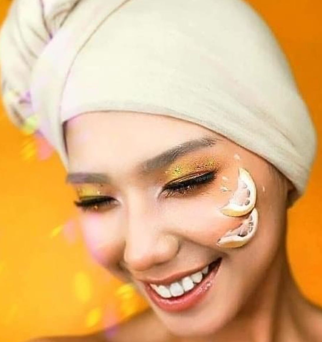 Smiling model wearing a cream towel head wrap against a vivid orange backdrop, with gold glitter eyeshadow and a citrus slice held at the cheek