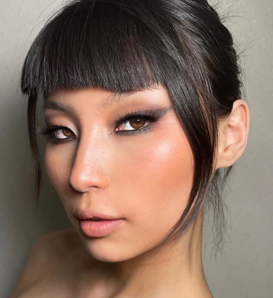 Beauty close-up with blunt micro-fringe bangs, sleek hair, a defined soft smoky brown eye and glowing peach-toned skin