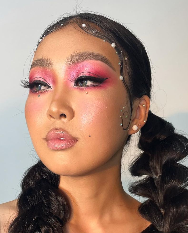 Model with two braided pigtails, intricate pink and magenta eyeshadow, and small rhinestone gem accents scattered across the cheeks and temples