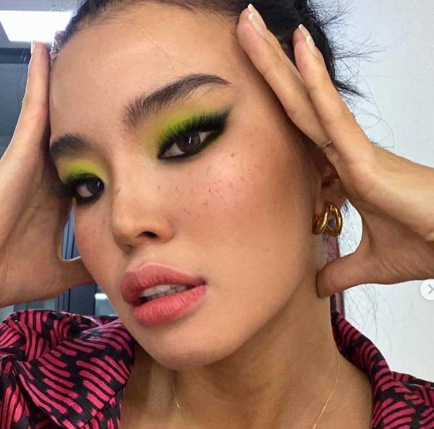 Energetic close-up with hands framing the face, bold lime-green and yellow eyeshadow blended with black liner, gold hoop earrings, and a pink animal-print top