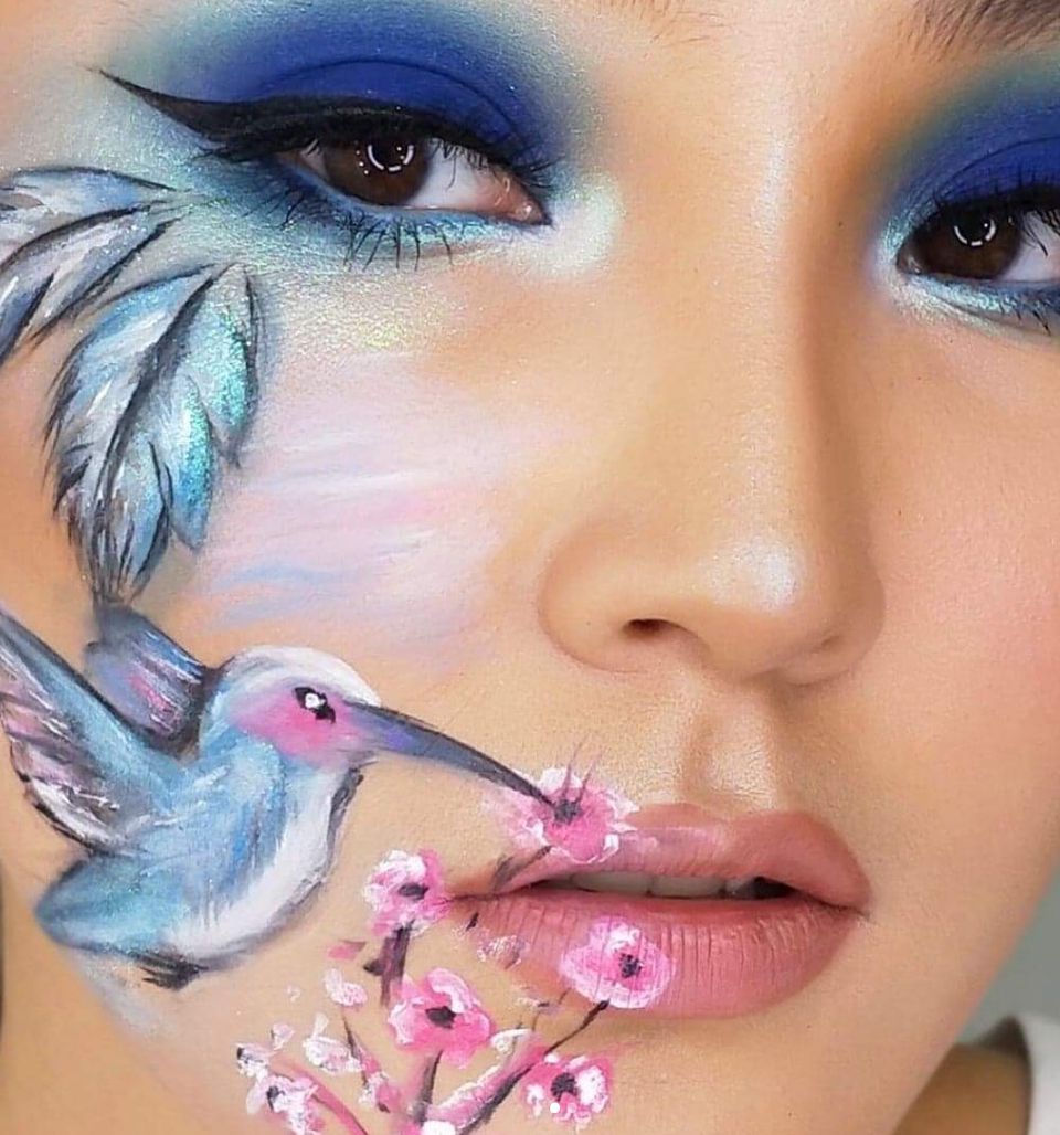 Extreme close-up of hand-painted face art depicting a hummingbird and cherry-blossom branch on the cheek, paired with a deep blue smoky winged eye